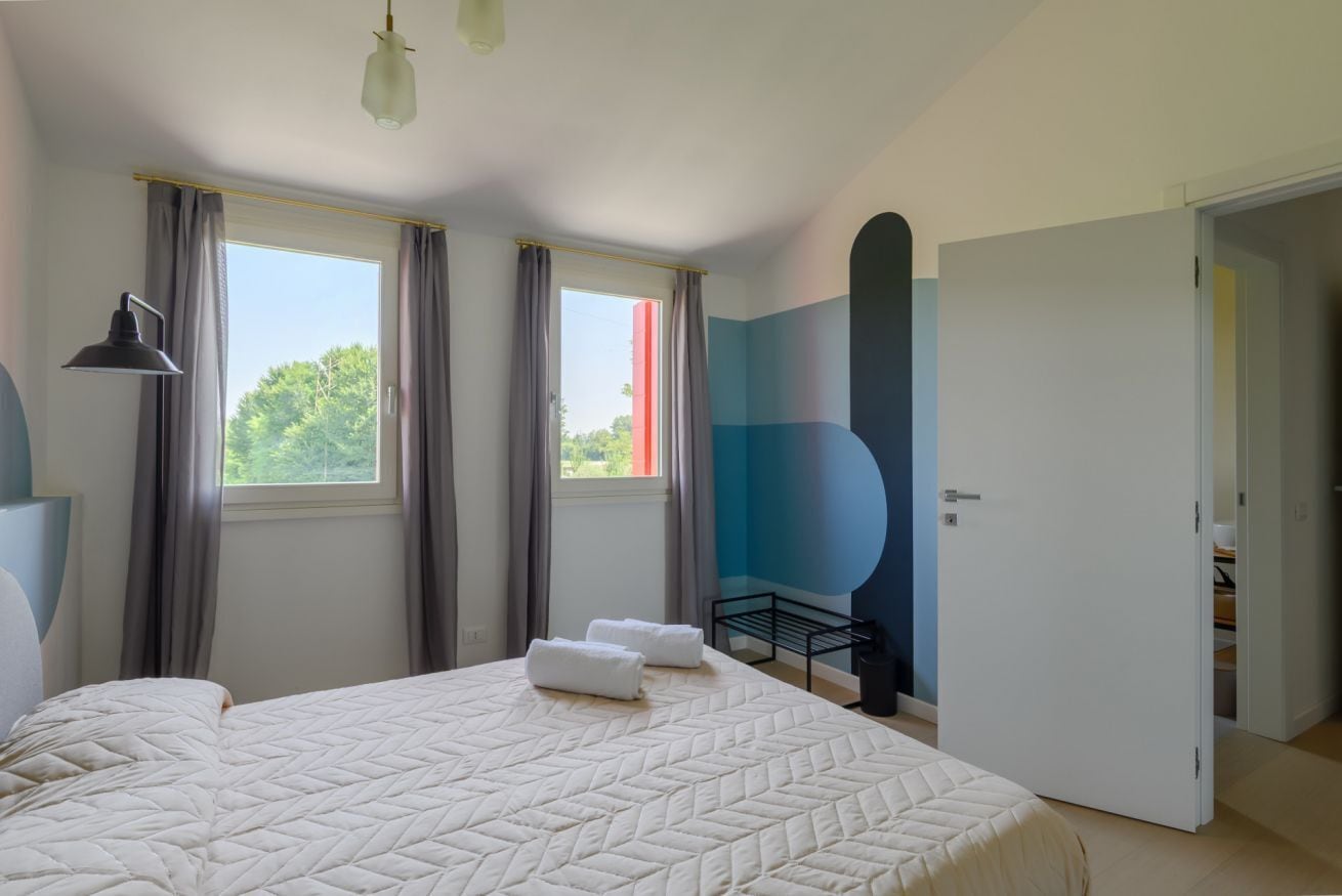 A bedroom with a large bed, two windows with curtains, a bedside lamp, and a door leading to another room.