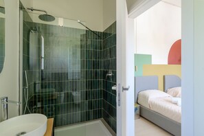 Een moderne badkamer met een douchecabine van glas, een witte wastafel en een bed met een grijs hoofdeinde.