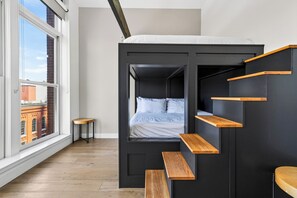 A bunk bed with wooden shelves, a small table, and a view of a brick building through a large window. - Group-Friendly Condo + King Suite | Steps to Music Hall + Broadway | Mic Drop (Nashville)