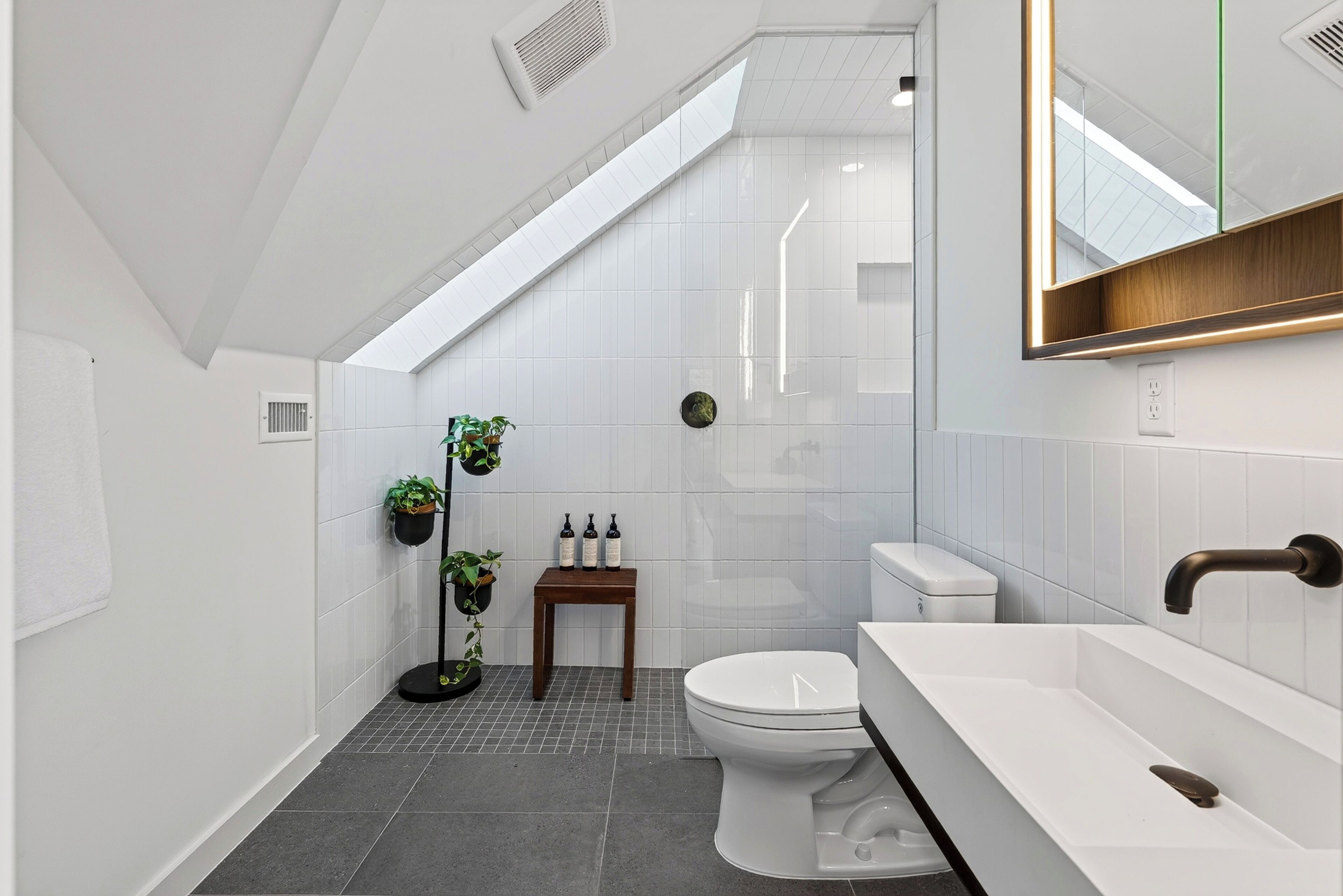 Een kleine badkamer met dakraam, witte tegels, toilet, wastafel en een klein houten tafeltje met planten.