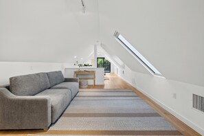 Et moderne loftværelse med skrå vægge, en grå sofa, et lille skrivebord og udsigt gennem et ovenlys.
