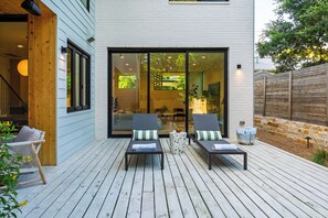 A modern outdoor deck with two lounge chairs, a small table, and a glass door leading to an interior space. - Plunge Pool + Firepit + Skyline Deck | Steps to SoCo | Elizabeth House (Austin)