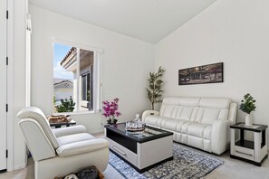 A living room with a white sofa, a white armchair, a glass coffee table, a small side table, and a painting on the wall. - Golf Views + BBQ Patio | Mins to Indian Wells | Palm & Par (Palm Desert)