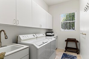 A compact laundry room with a front-loading washer and dryer, a sink, and a window with a view of greenery. - Golf Views + BBQ Patio | Mins to Indian Wells | Palm & Par (Palm Desert)