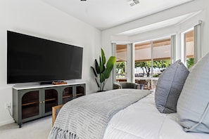 A modern bedroom with a large flat-screen TV, a bed with white and gray bedding, and a view of the outdoors through large windows. - Golf Views + BBQ Patio | Mins to Indian Wells | Palm & Par (Palm Desert)