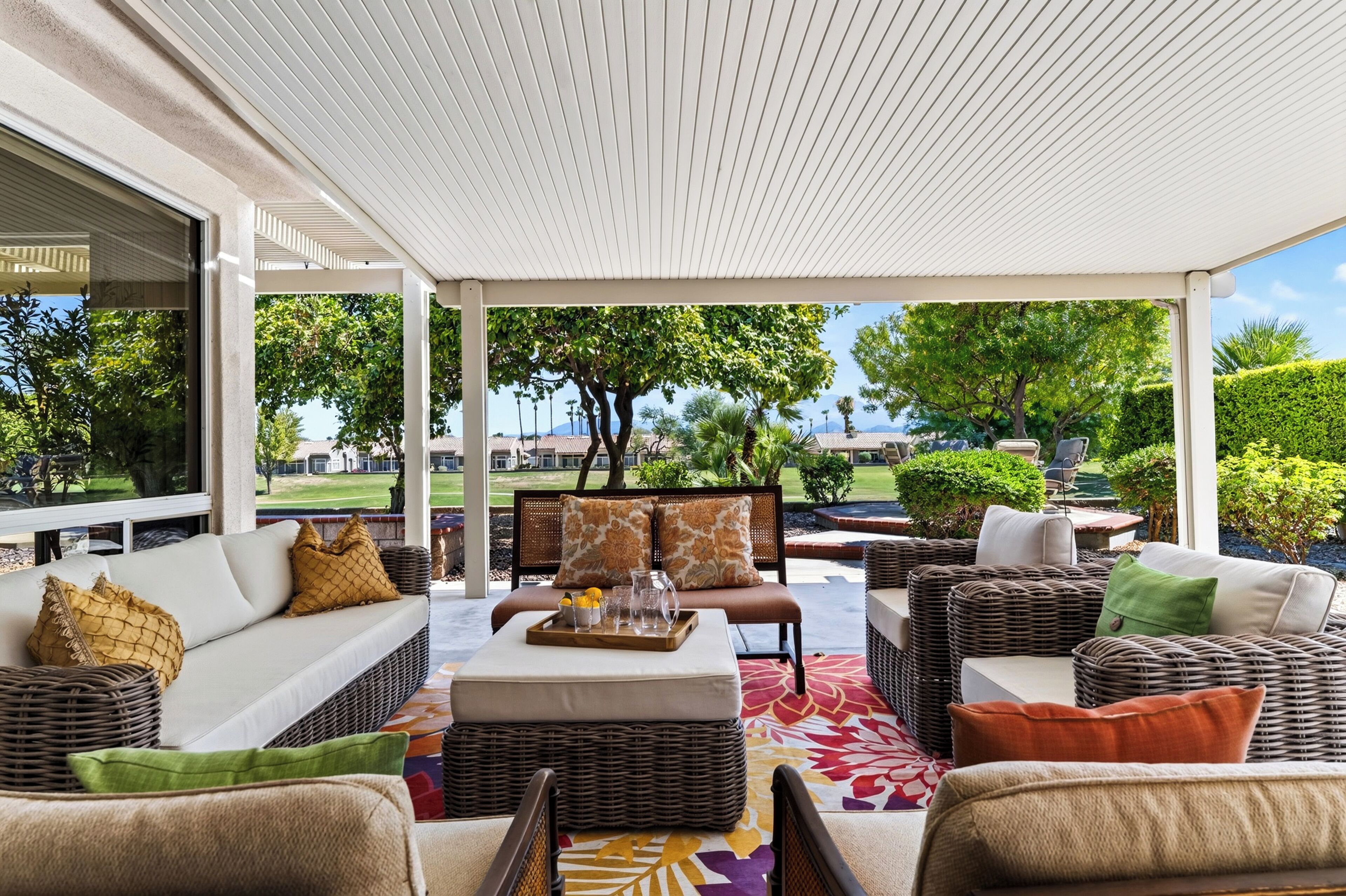 A covered outdoor seating area with wicker furniture, a coffee table with drinks, and a view of a residential neighborhood.