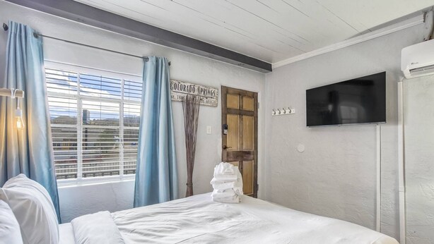 Ein Schlafzimmer mit einem Bett, einem Fenster mit Vorhängen, einer Holztür und einem an der Wand befestigten Fernseher.