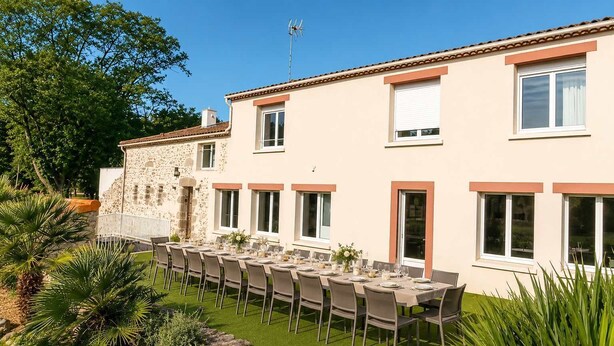 A building with a stone facade and a flat roof, a long dining table set for a meal, and a well-maintained lawn.
