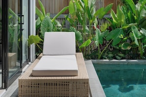 Una silla de descanso junto a la piscina, con cojín, ubicada al lado de un ventanal de vidrio, con una piscina y plantas tropicales al fondo.