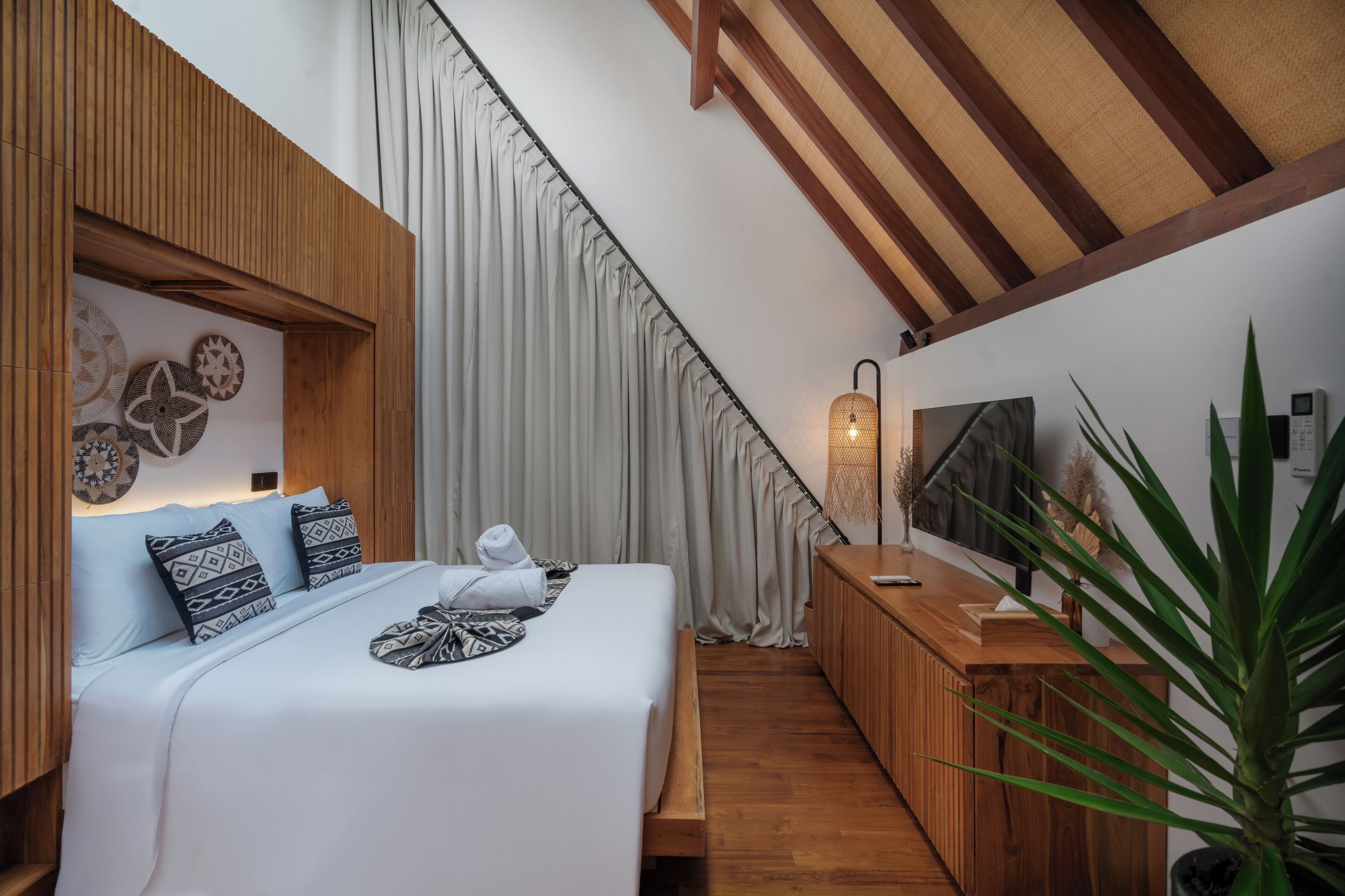 A modern hotel room with a bed, wooden paneling, a large window with curtains, a wooden desk, a plant, and a wall-mounted TV.
