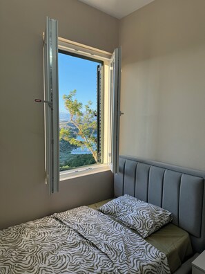 A room with a bed, a window with open shutters, and a view of greenery outside.