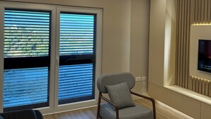 A modern living room with a grey armchair, a black sofa, and a large window with blinds. - The Lake Villa Tirana (Tiranë)