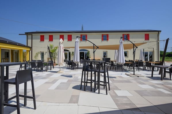 An outdoor seating area with tables and chairs, a building with red shutters, and a clear sky.