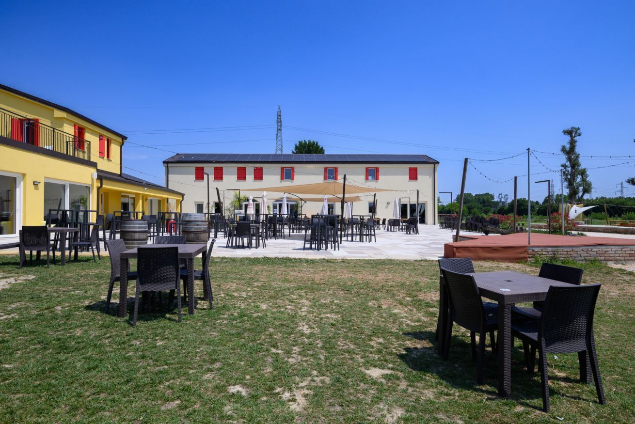 Ein Außenbereich mit Tischen und Stühlen, eine überdachte Terrasse und ein Gebäude mit roten Fensterrahmen.