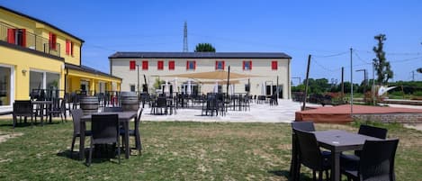 An outdoor seating area with tables and chairs, a covered patio, and a building with red window frames.