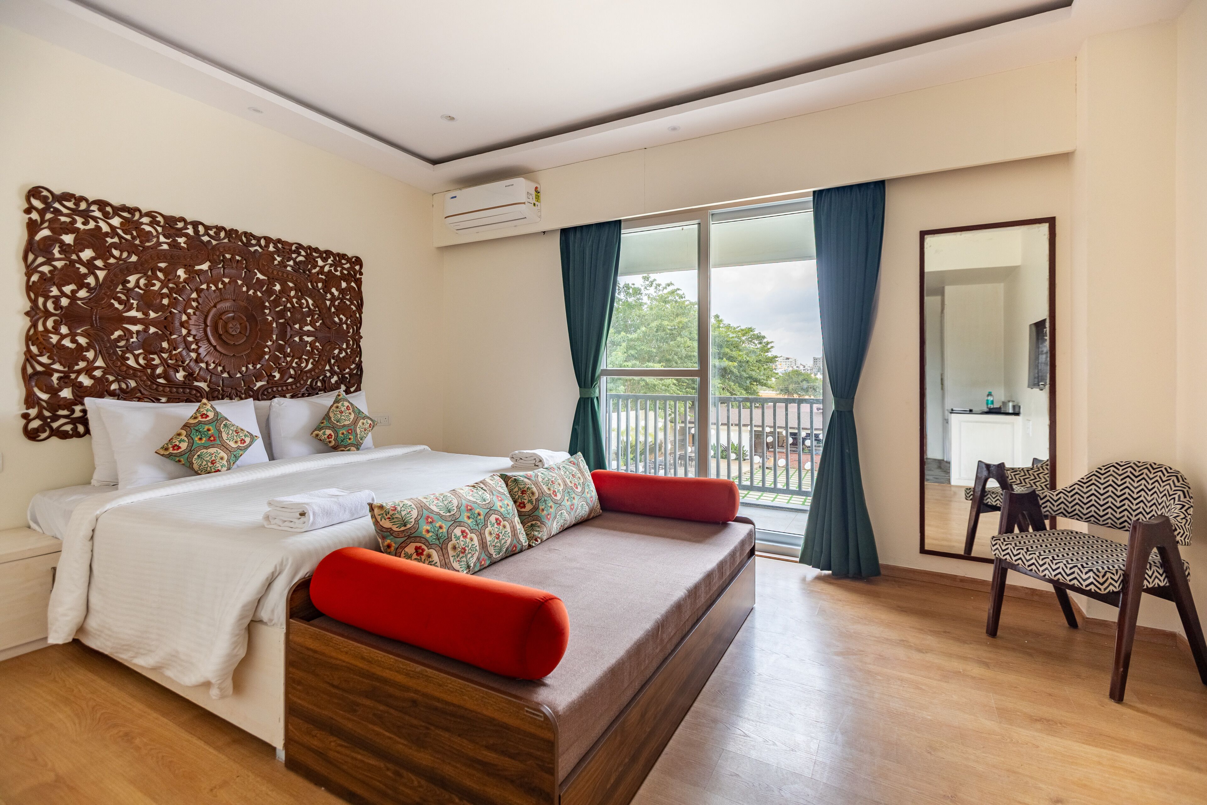 A hotel room with a bed, a wooden headboard, a balcony with curtains, a chair, and a large mirror.