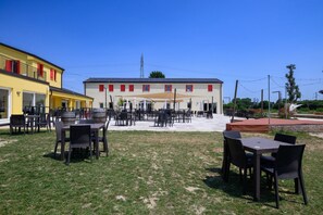 Ein Außenbereich mit Tischen und Stühlen, eine überdachte Terrasse und ein Gebäude mit roten Fensterrahmen.