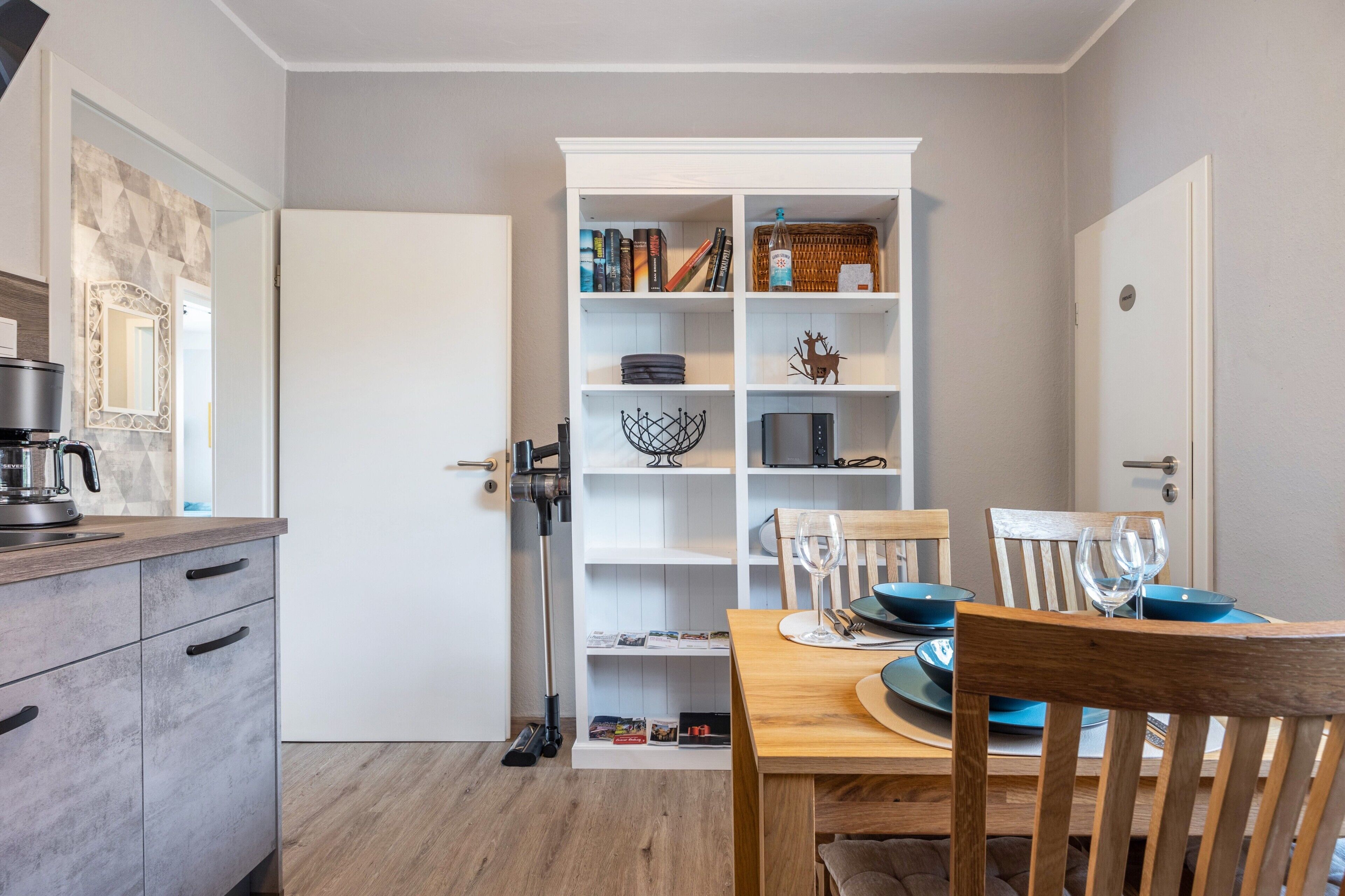 Eine moderne Küche mit Essbereich, mit einem weißen Schrank, der mit Büchern und Dekorationen ausgestattet ist, einer Kaffeemaschine und einem für zwei Personen gedecktem Esstisch.