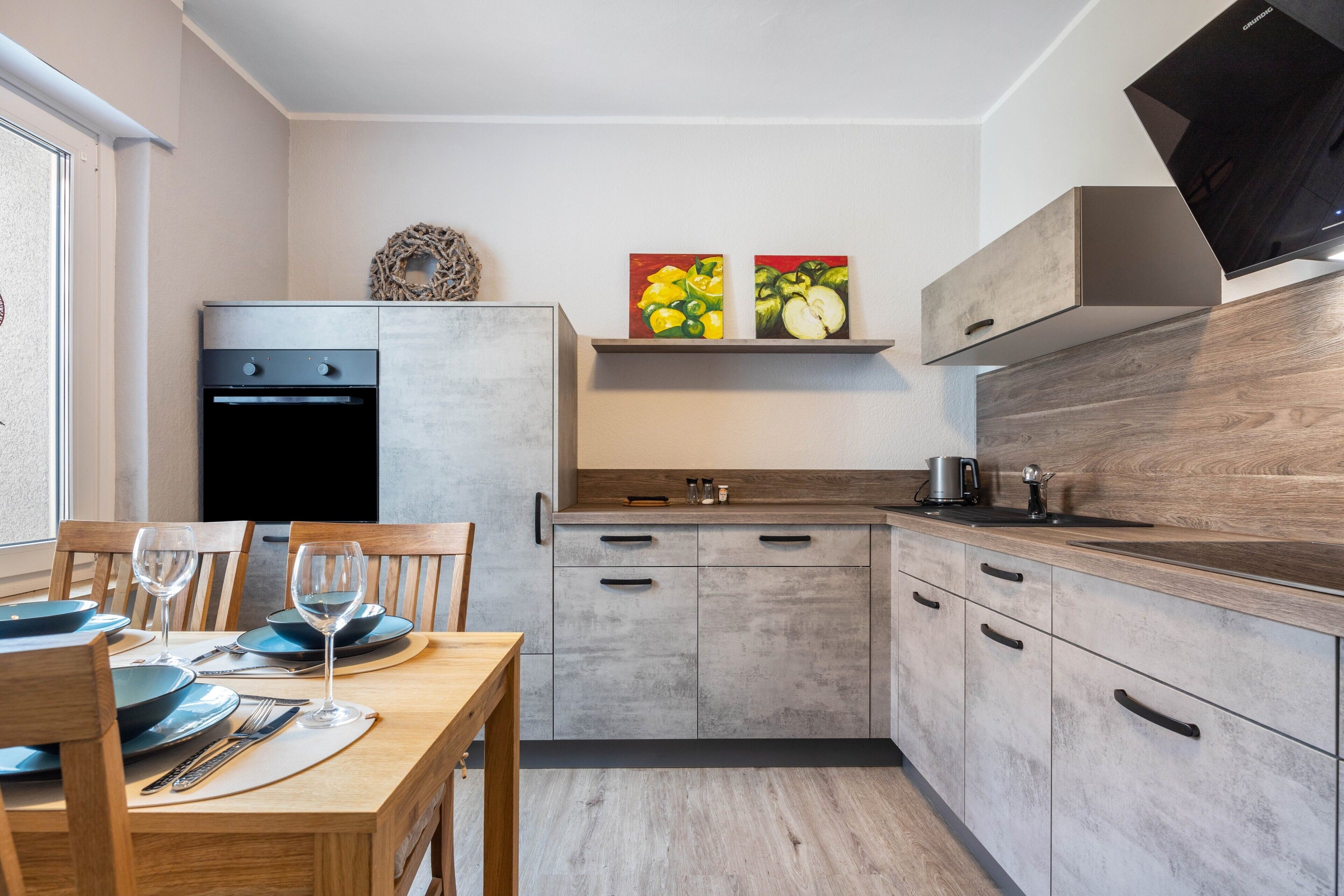 A modern kitchen with built-in appliances, a dining area with a wooden table and chairs, and a wall-mounted shelf with artwork.