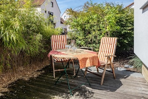 Eine Außenpatio mit einem Holztisch und zwei gestreiften Stühlen, umgeben von üppiger Bepflanzung und einem weißen Gebäude im Hintergrund.