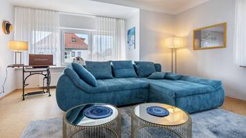 En moderne stue med en hjørnesofa, to runde sofaborde, en lampe og udsigt til en bygning gennem store vinduer.