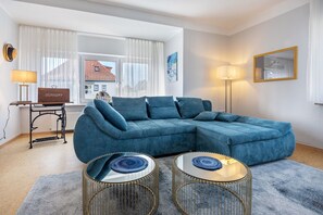 Una sala moderna con un sofá modular, dos mesitas redondas, una lámpara y vista a un edificio a través de amplios ventanales.