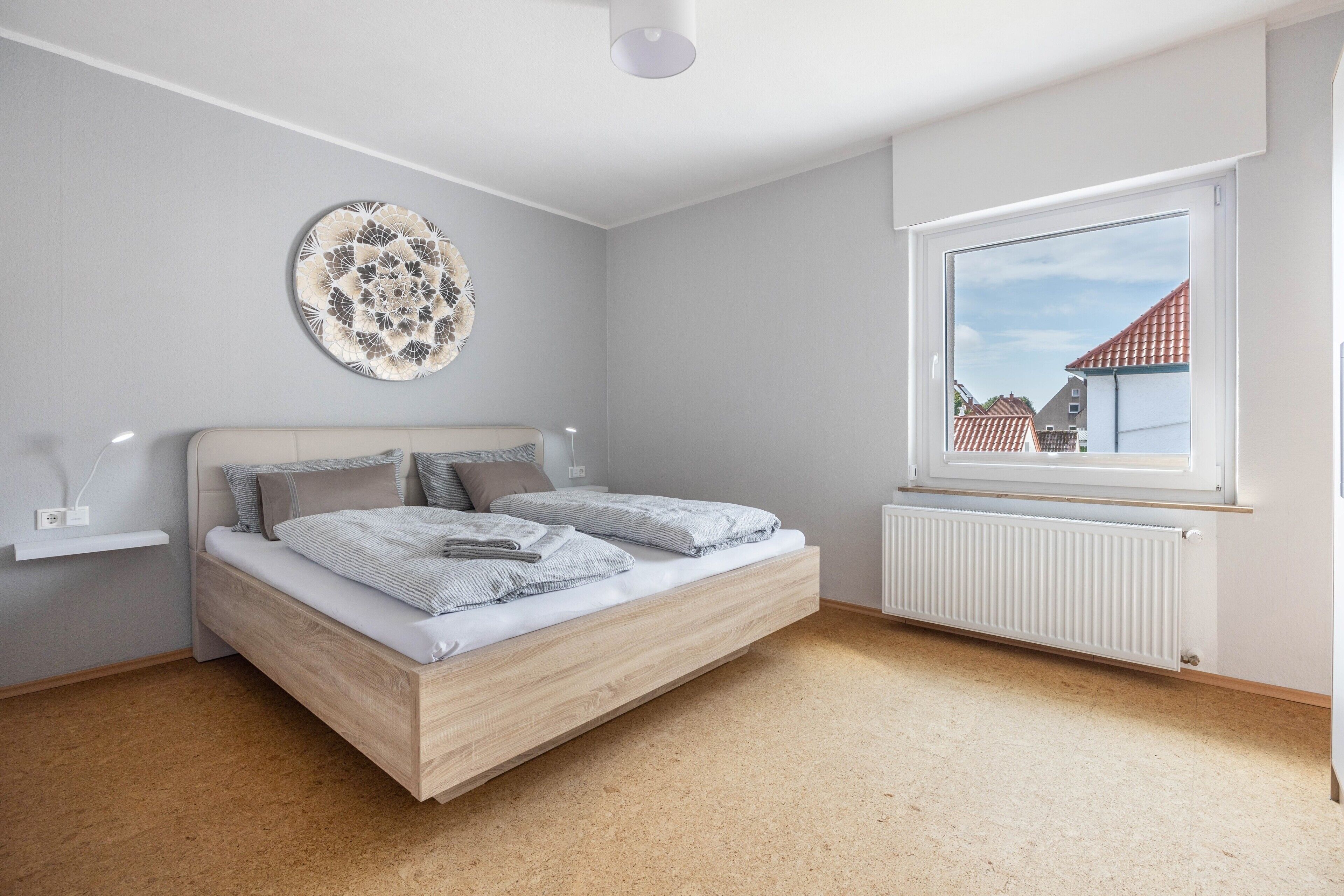 A bedroom with a bed, a wall-mounted decorative plate, a window with a view of buildings, and a radiator.