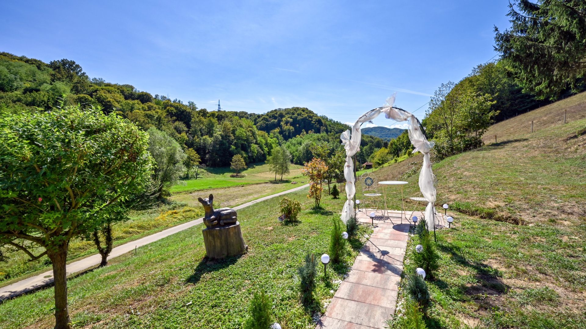 A pathway leading to a white archway with a statue on the left, surrounded by greenery and a clear sky.