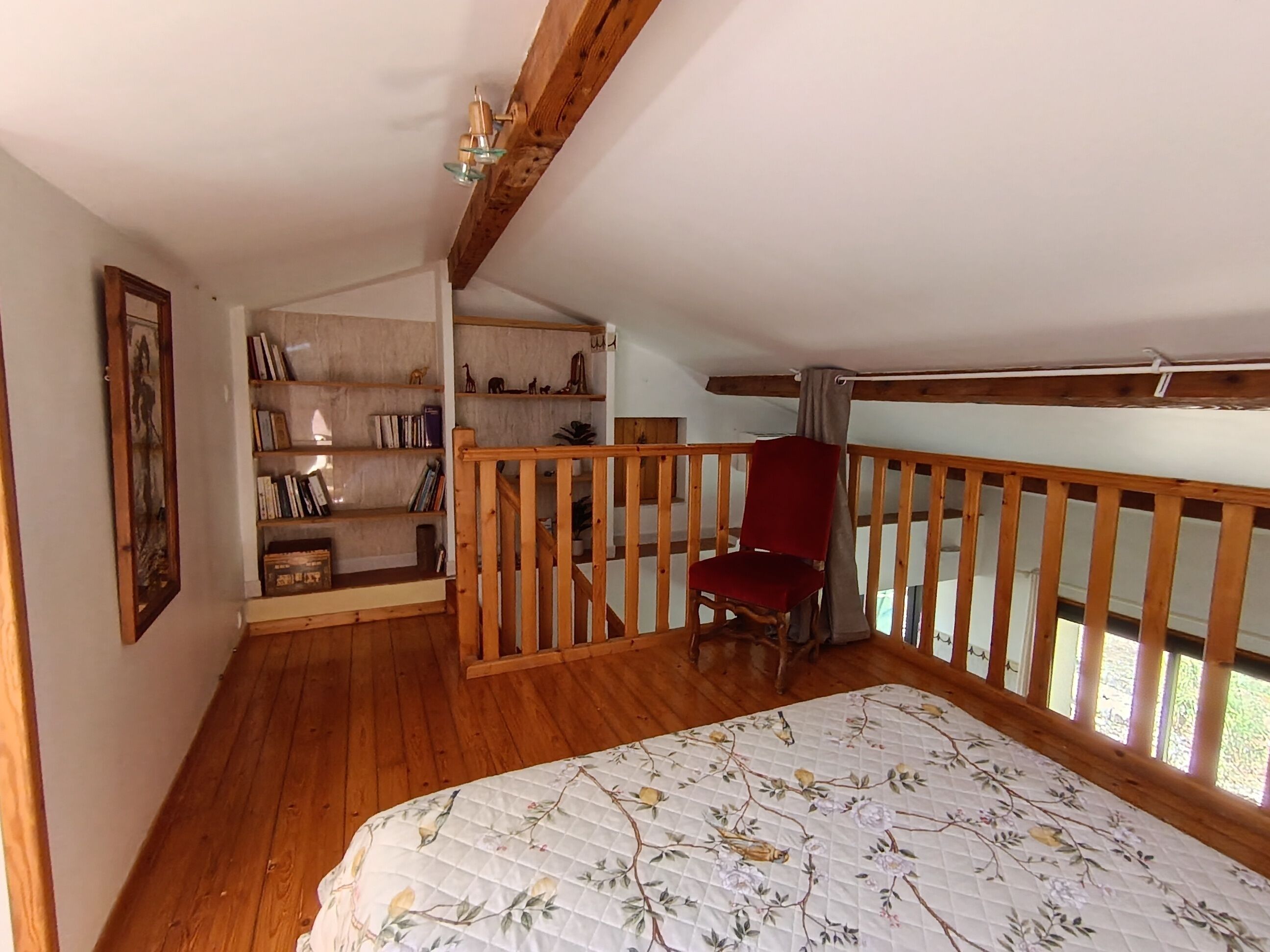 Una habitación con piso de madera, una silla roja, una cama con colcha floral, una estantería y una barandilla de madera.