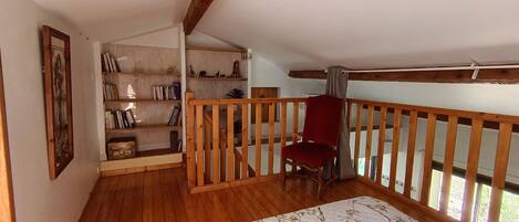 Una habitación con piso de madera, una silla roja, una cama con colcha floral, una estantería y una barandilla de madera.