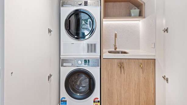Een kleine wasruimte met een voorlader wasmachine en droger, een wastafel met gouden kraan en houten kastjes.