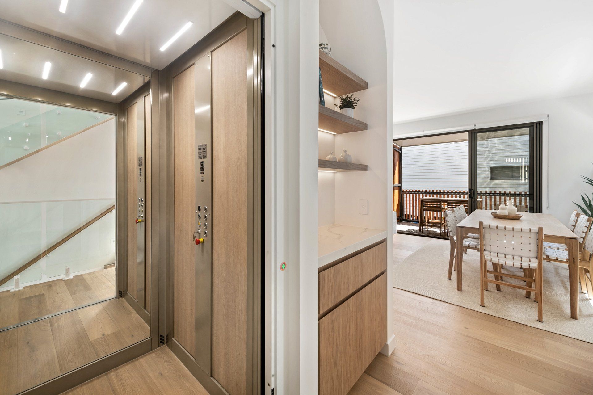 Un interno moderno con una scala in legno, una zona pranzo con tavolo e sedie, e vista all'esterno tramite una porta in vetro.