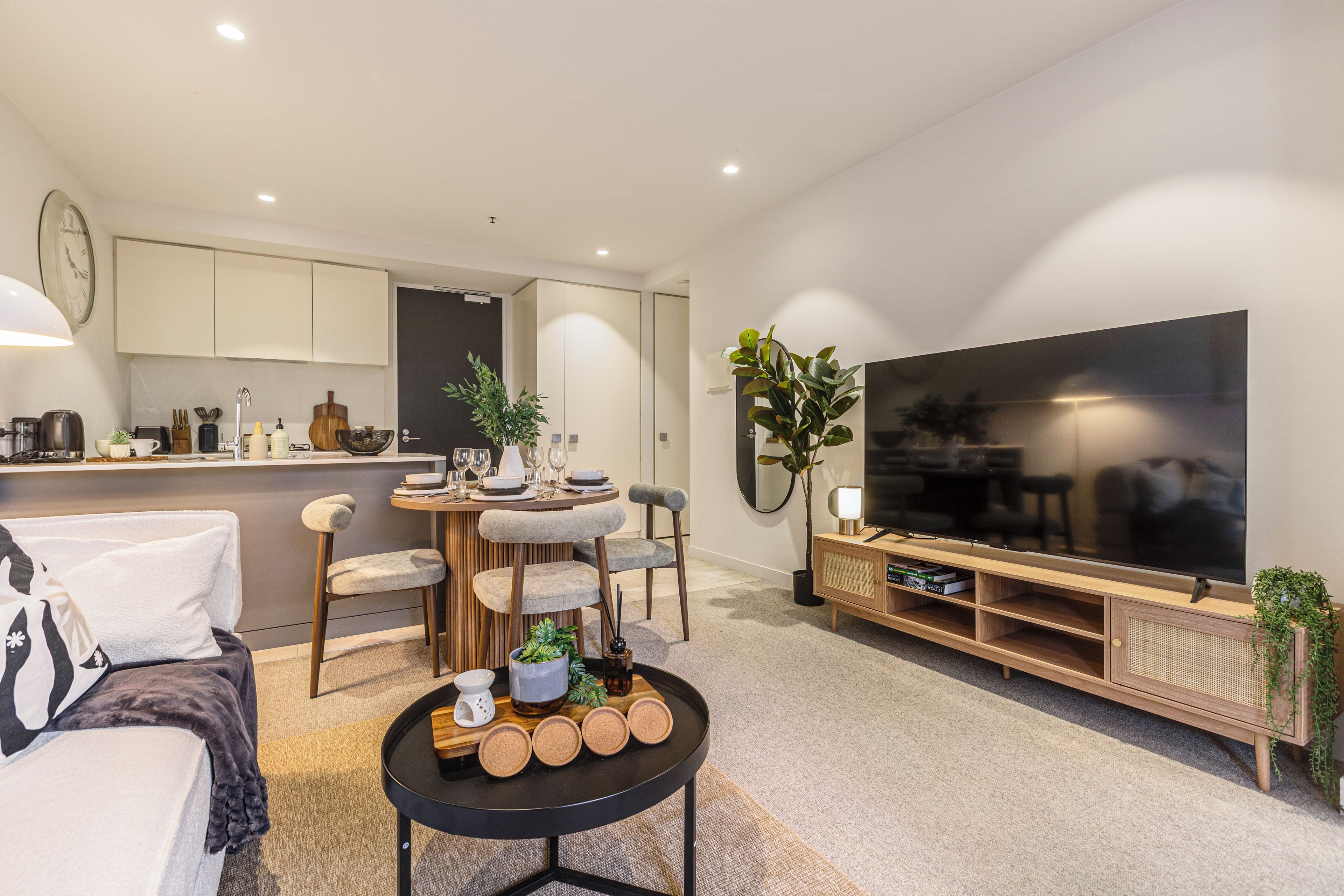 A modern living room with a flat-screen TV, a dining area with a round table, and a kitchen in the background.