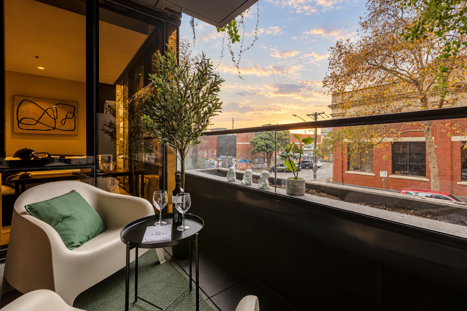 A balcony with a view of a cityscape, a table with a bottle and glasses, and a chair with a pillow.
