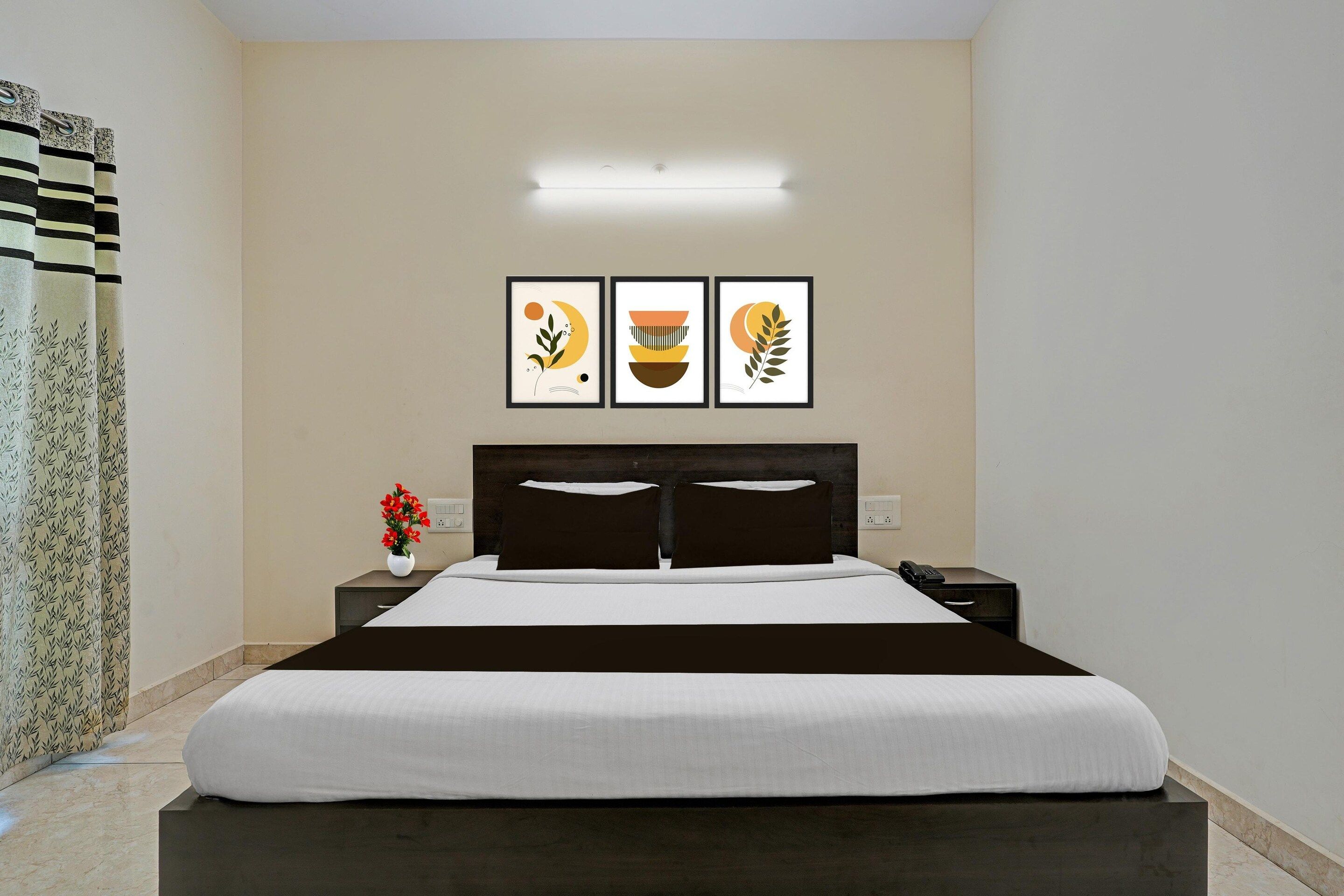A hotel room with a bed, a nightstand, a phone, and a wall-mounted light. There are three framed pictures on the wall.