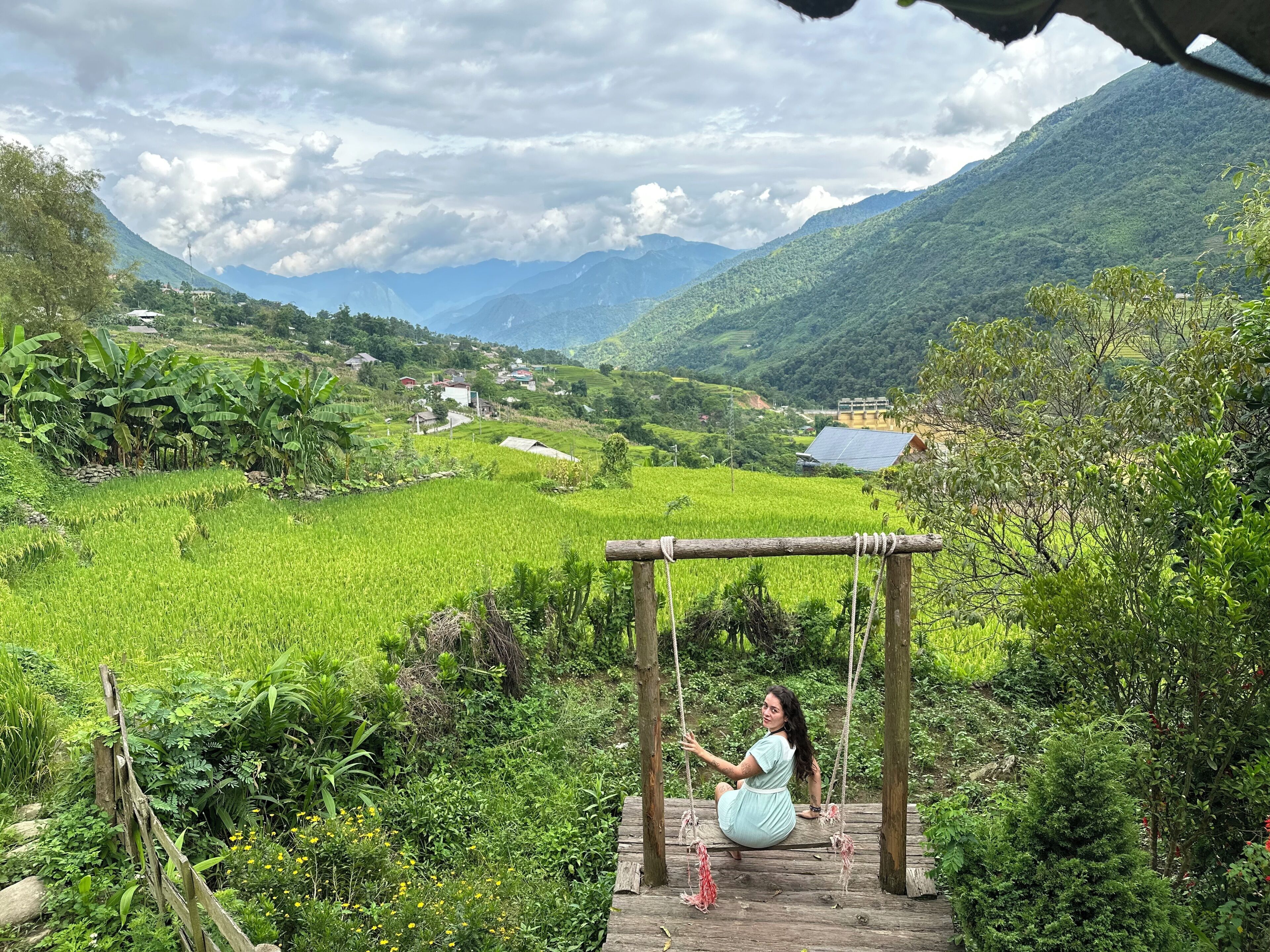 Une personne assise sur une balançoire, dans un paysage verdoyant et luxuriant, avec des montagnes en arrière-plan.