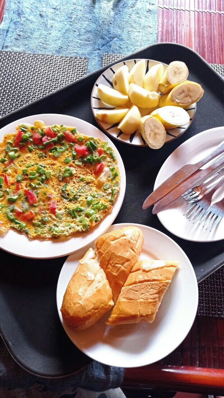 Una bandeja con una fuente de plátanos en rodajas, una fuente de huevos revueltos con vegetales, y una fuente de croissants.