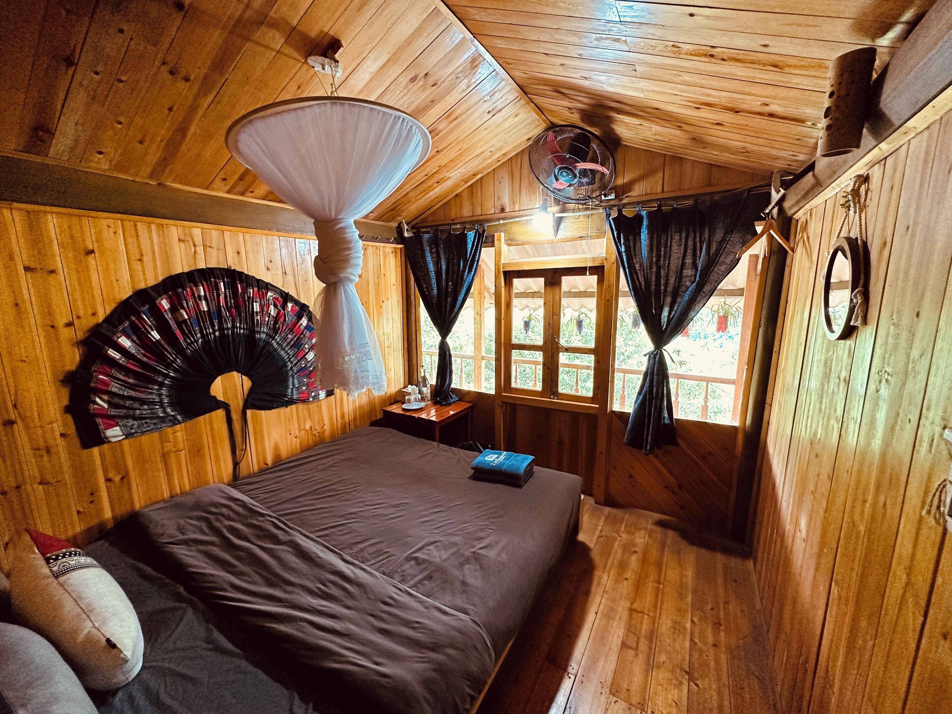A wooden cabin room with a bed, a fan, a window with curtains, and a hanging lamp.
