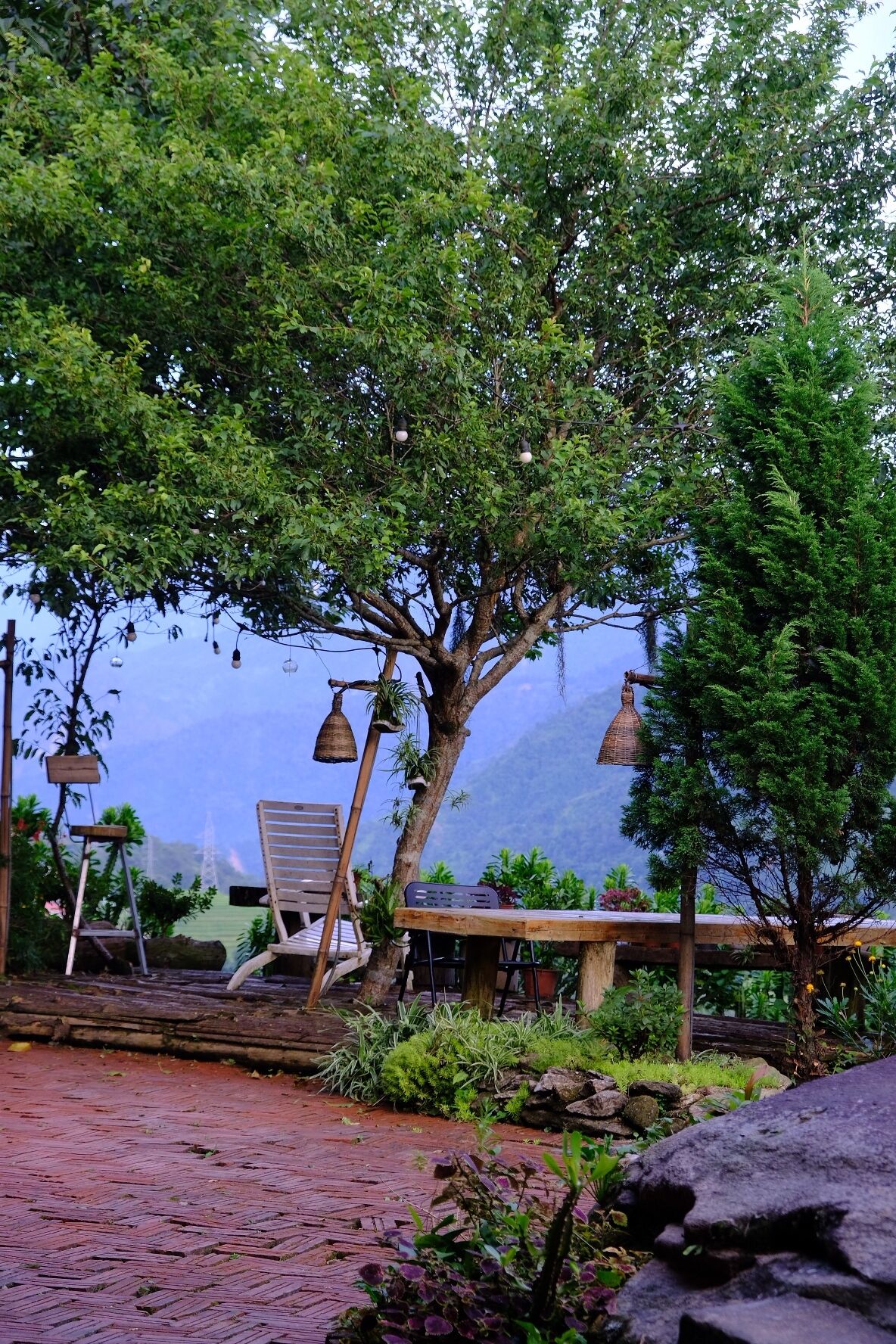 A serene outdoor seating area with a wooden table and chairs, surrounded by lush greenery and a tree.