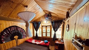 Una habitación de cabaña de madera con una cama, un ventilador, una mesa con una silla y una ventana con cortinas.