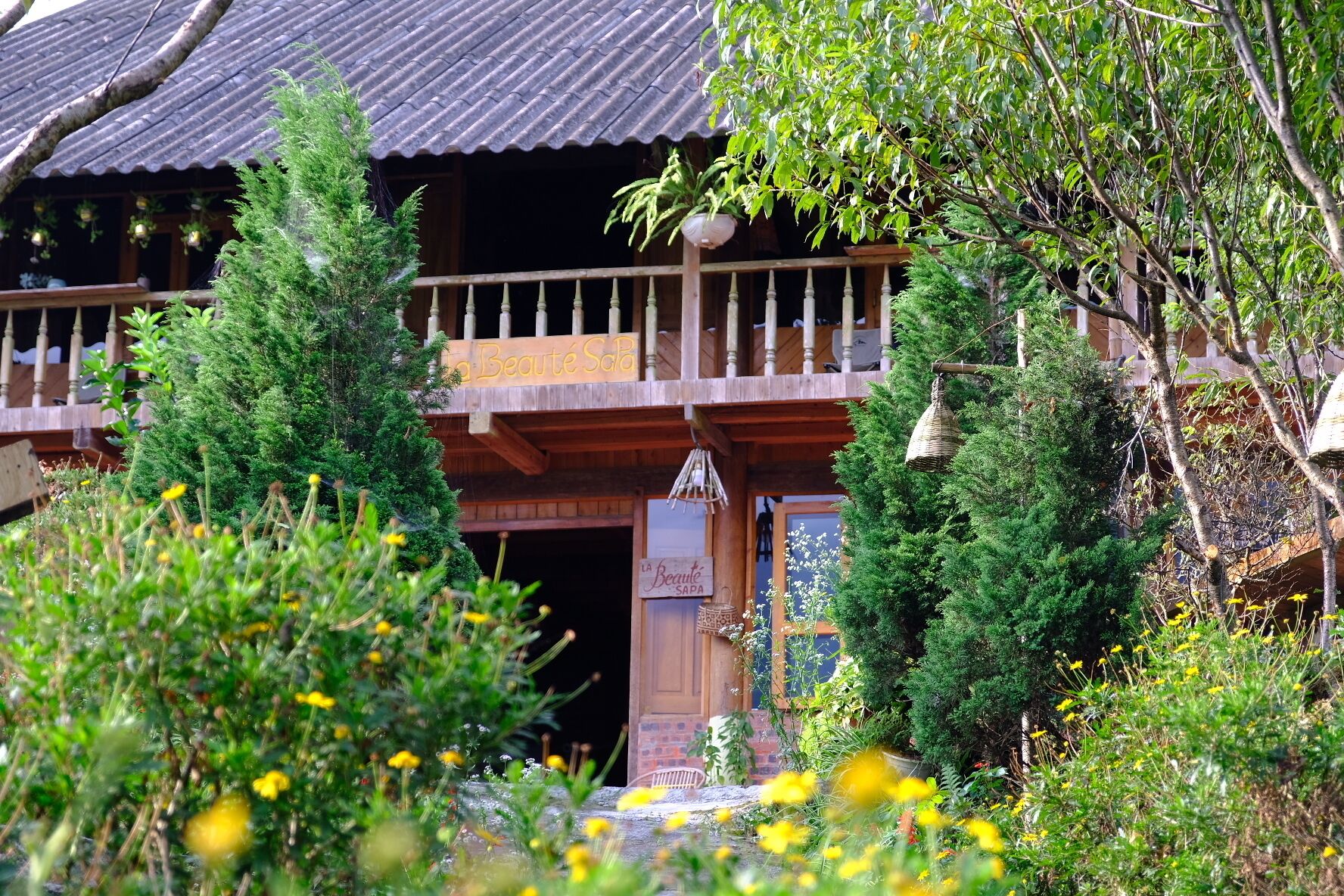 A traditional wooden building with a balcony, surrounded by lush greenery and yellow flowers.