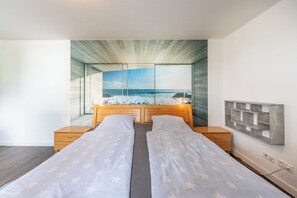 Une chambre avec une grande fenêtre, un lit avec deux oreillers, une tête de lit en bois et une table de chevet.