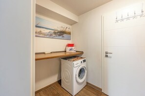 A laundry room with a washing machine, a shelf with a red container, and a painting of a beach scene. - "Landlust" Vacation Rental in Husum-Schauendahl for up to 4 people (Husum-Schauendahl)