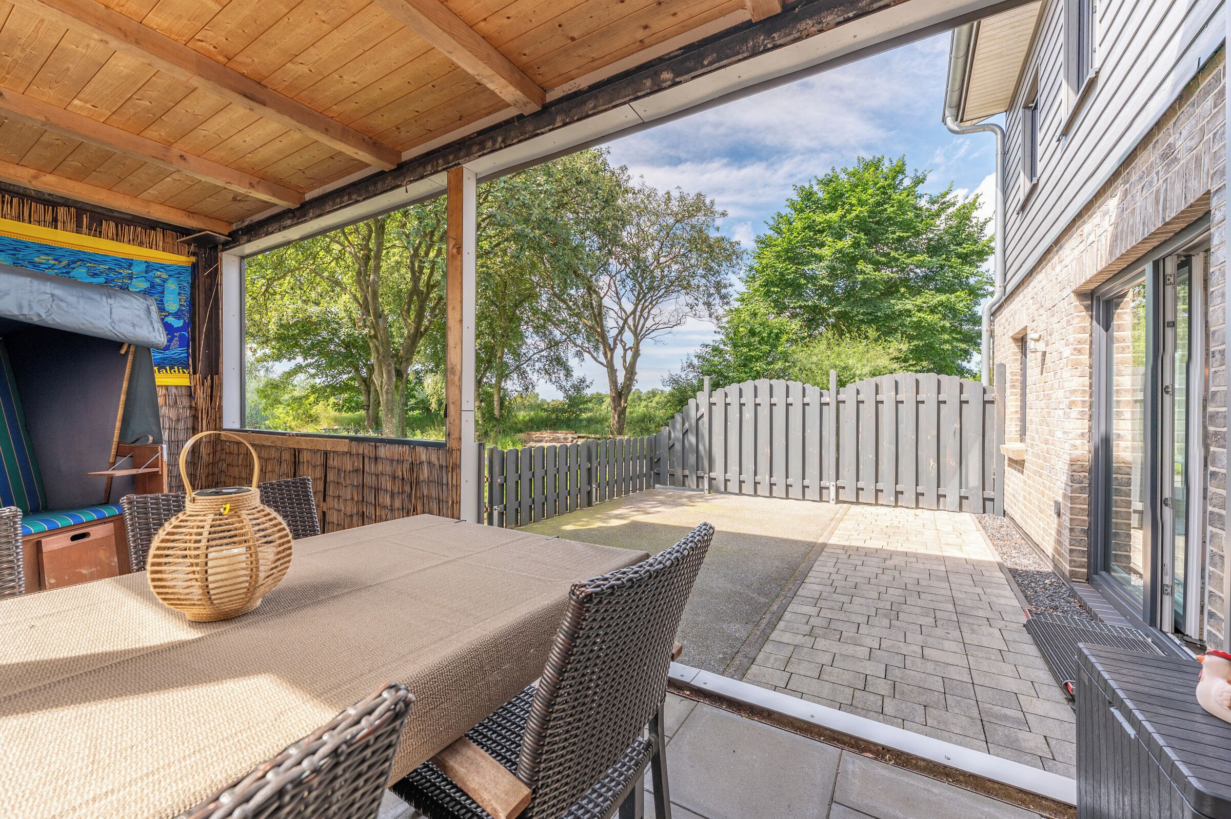 Eine Terrasse im Freien mit Tisch und Stühlen, einem Korbwäschesessel und Blick auf ein Ziegelgebäude und Bäume.