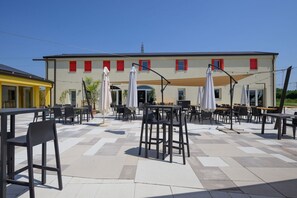 Ein Sitzbereich im Freien mit Tischen und Stühlen, ein Gebäude mit roten Fensterläden im Hintergrund und ein klarer Himmel.