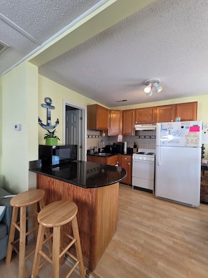 A kitchen with wooden cabinets, a black countertop, a white refrigerator, and a wooden island with stools. - Beach Breeze Bungalow | Outdoor Oasis | 2BR/2BA (Myrtle Beach)