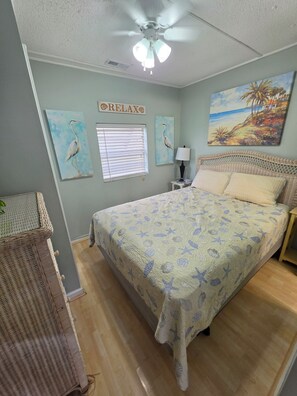 A bedroom with a bed, a ceiling fan, a window with blinds, and a painting on the wall. - Beach Breeze Bungalow | Outdoor Oasis | 2BR/2BA (Myrtle Beach)