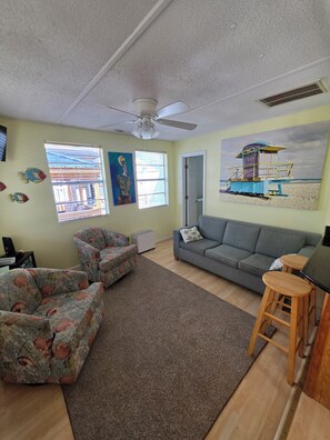 A living room with a grey sofa, floral armchairs, a ceiling fan, and a beach-themed painting. - Beach Breeze Bungalow | Outdoor Oasis | 2BR/2BA (Myrtle Beach)