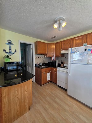 A kitchen with wooden cabinets, a black countertop, a stove, and a refrigerator. - Beach Breeze Bungalow | Outdoor Oasis | 2BR/2BA (Myrtle Beach)