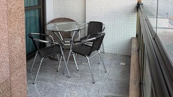 Uma varanda com mesa de vidro e cadeiras de vime, paredes com azulejos e janela com vista para a rua.